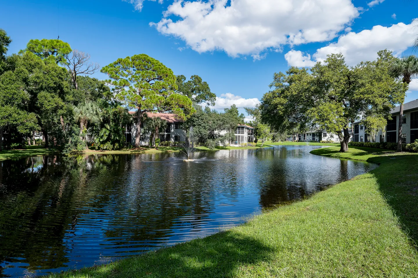 A serene pond with a central water fountain, bordered by grassy banks, leafy trees, and apartment buildings in the background under a blue sky.