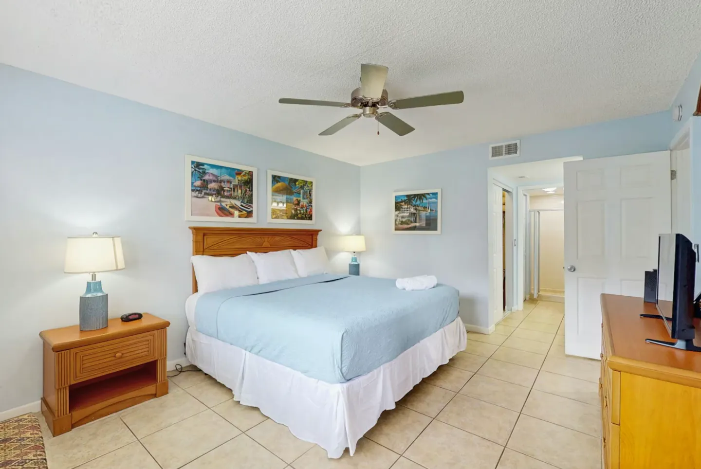 Bedroom with light blue walls, tiled floors, a wooden bed with light blue bedding, ceiling fan, and an open doorway leading to a bathroom.