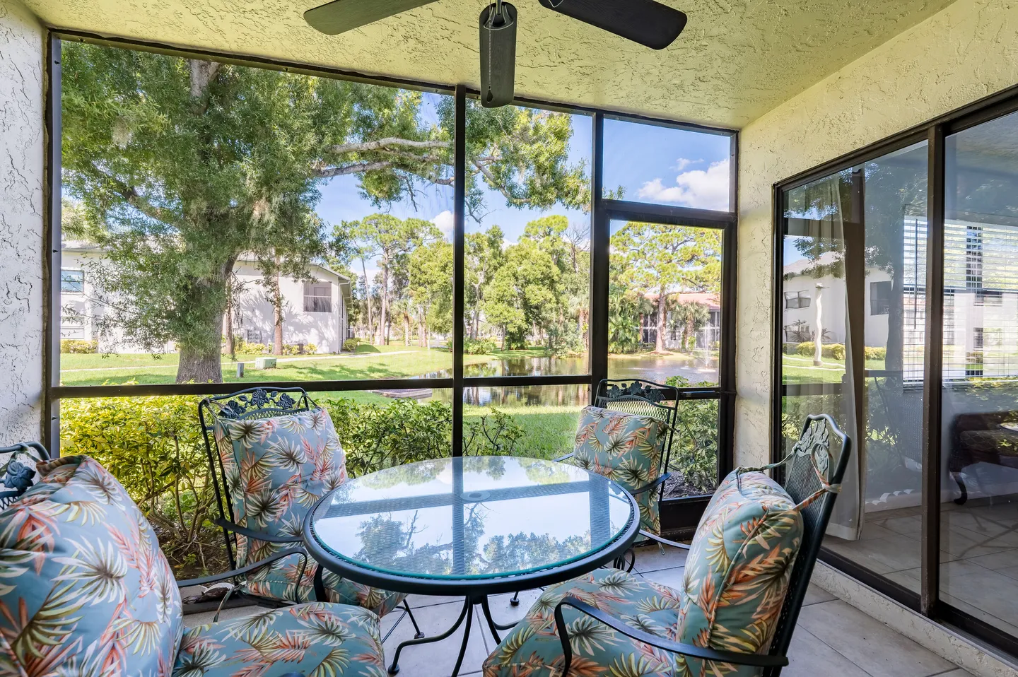 Screened porch furnished with a glass-top table, cushioned armchairs, and a ceiling fan, overlooking a pond and trees.