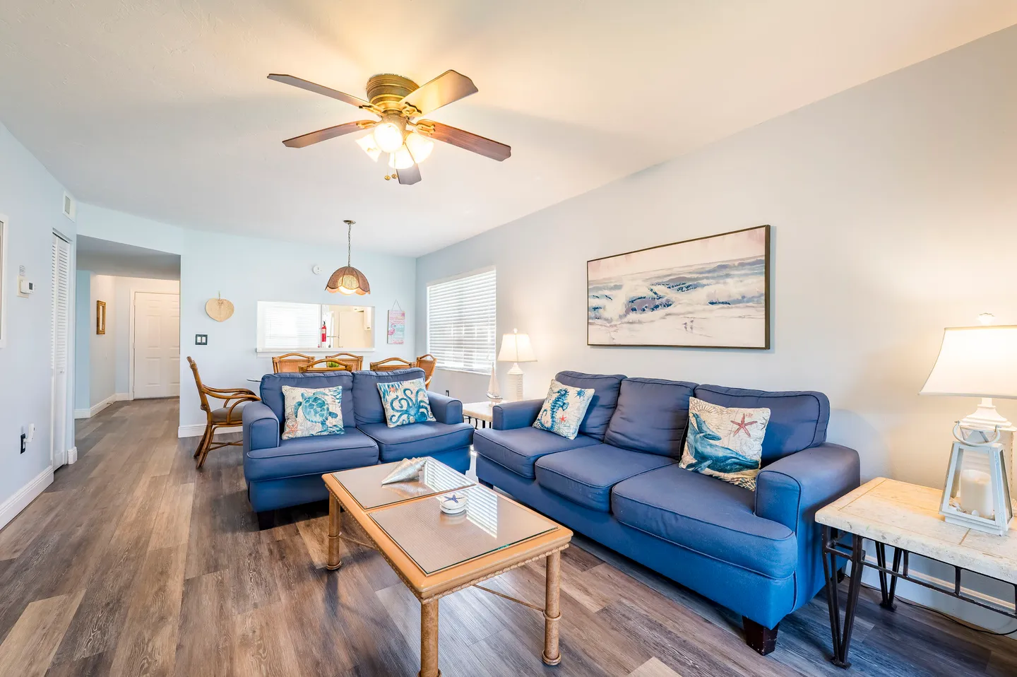 Living room with blue sofas, coffee table, wood-style flooring, and a ceiling fan, leading into a dining area.