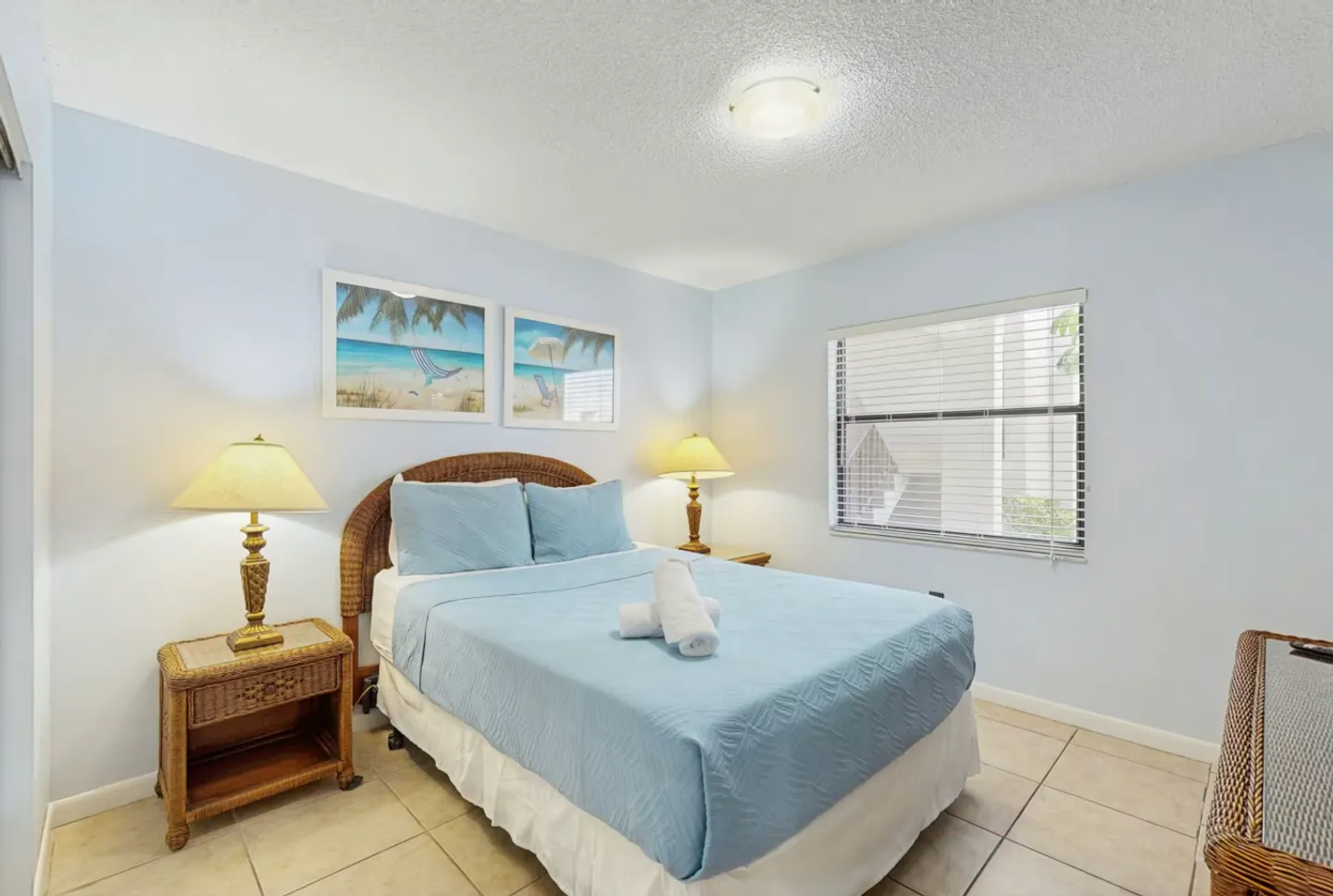 Bedroom with blue walls, tile flooring, a bed made with blue bedding, and wicker nightstands with lamps.