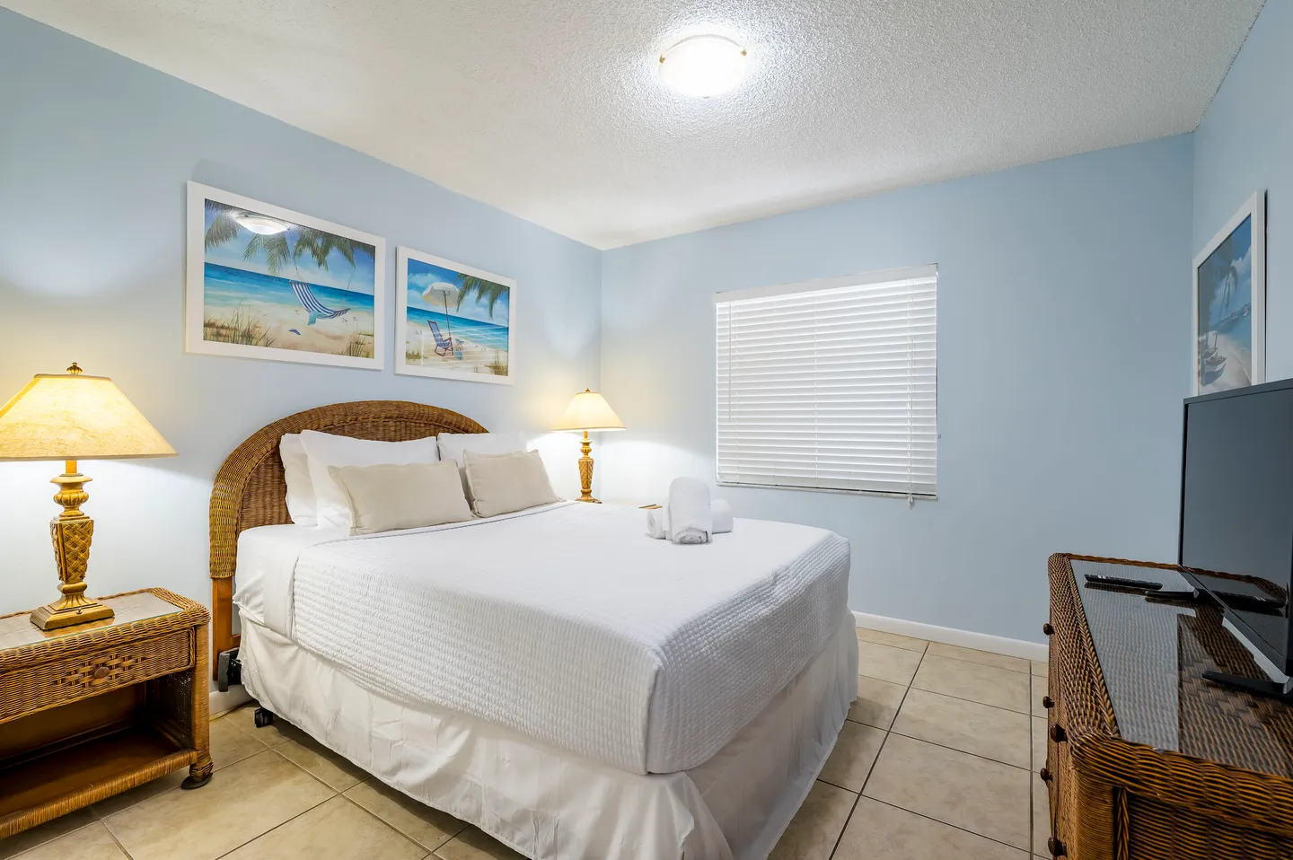 Bedroom with light blue walls, a bed with a wicker headboard, tiled floors, and a television on a wicker dresser.