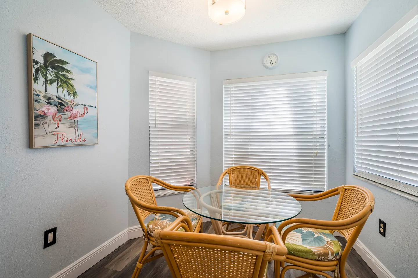 Breakfast nook with a round glass-top table, wicker armchairs, and windows with white blinds.