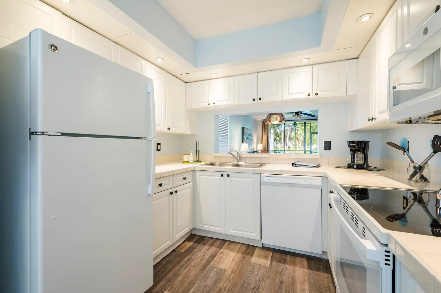 Kitchen with white cabinetry, white appliances, tiled countertops, wood-style flooring, and a passthrough opening into a living space.