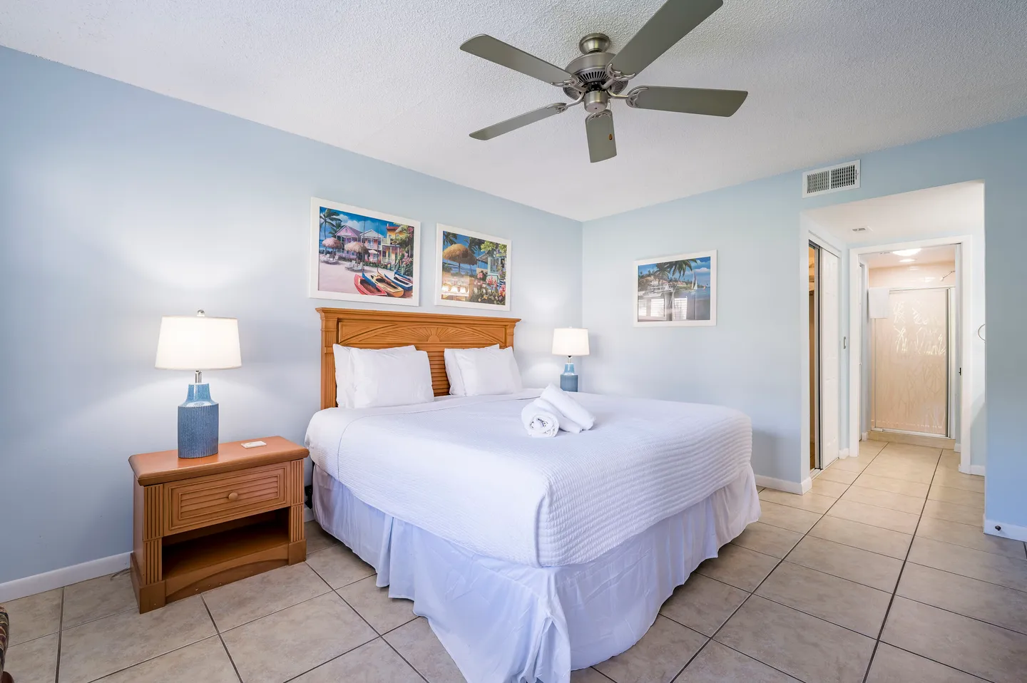 Bedroom with light blue walls, tiled floor, a bed with a wooden headboard and white linens, flanked by nightstands with lamps.