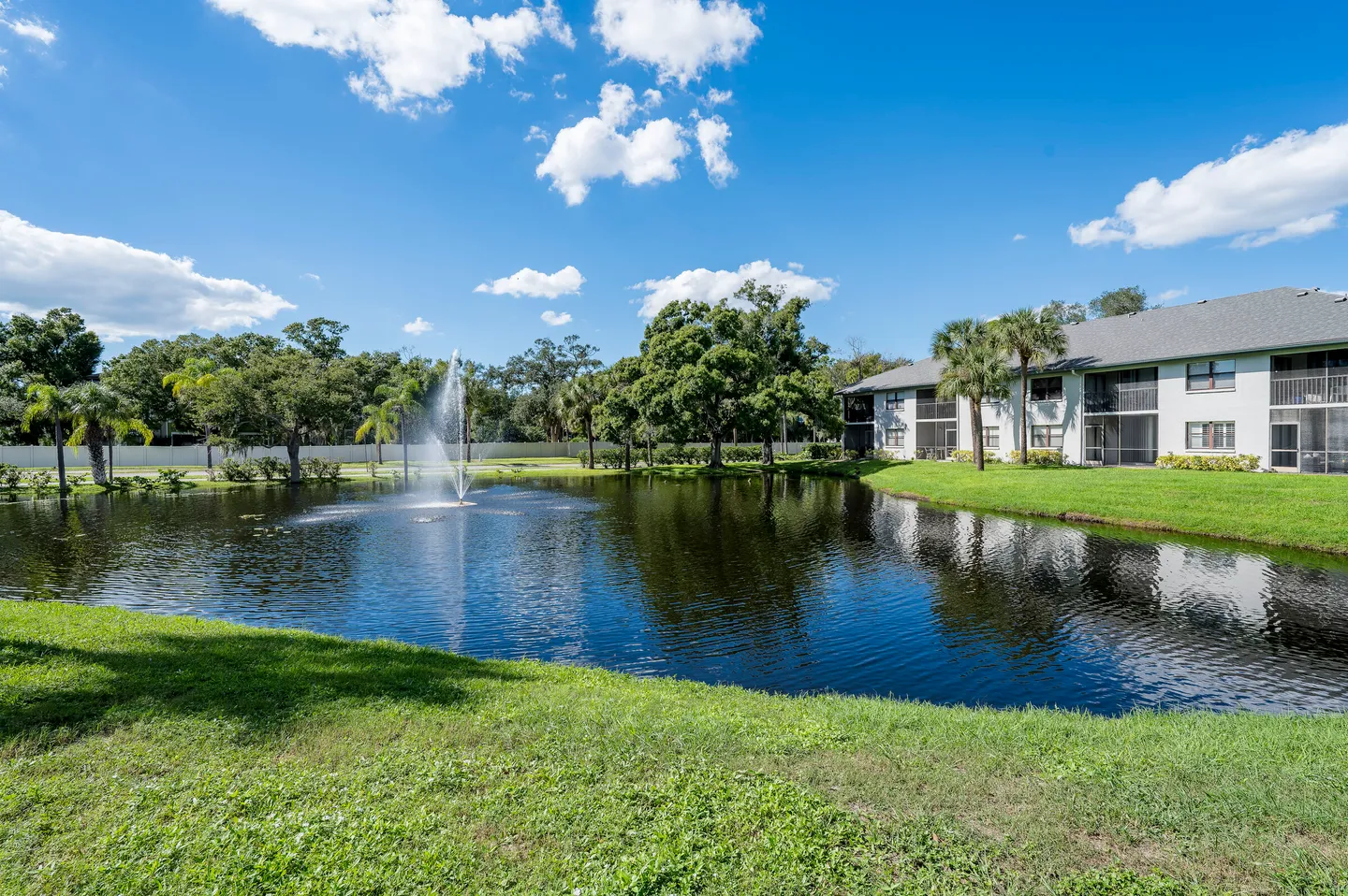 A water fountain spraying in a pond surrounded by green lawns, trees, and an apartment building under a blue sky.