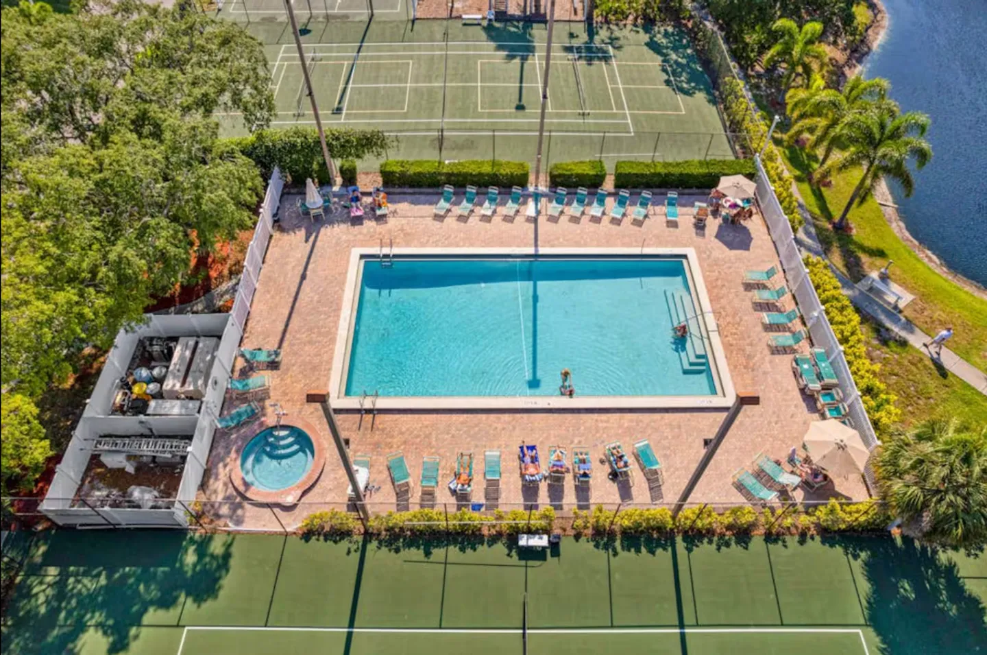 Aerial view of a community swimming pool, hot tub, and tennis courts surrounded by a paved deck and lounge chairs.