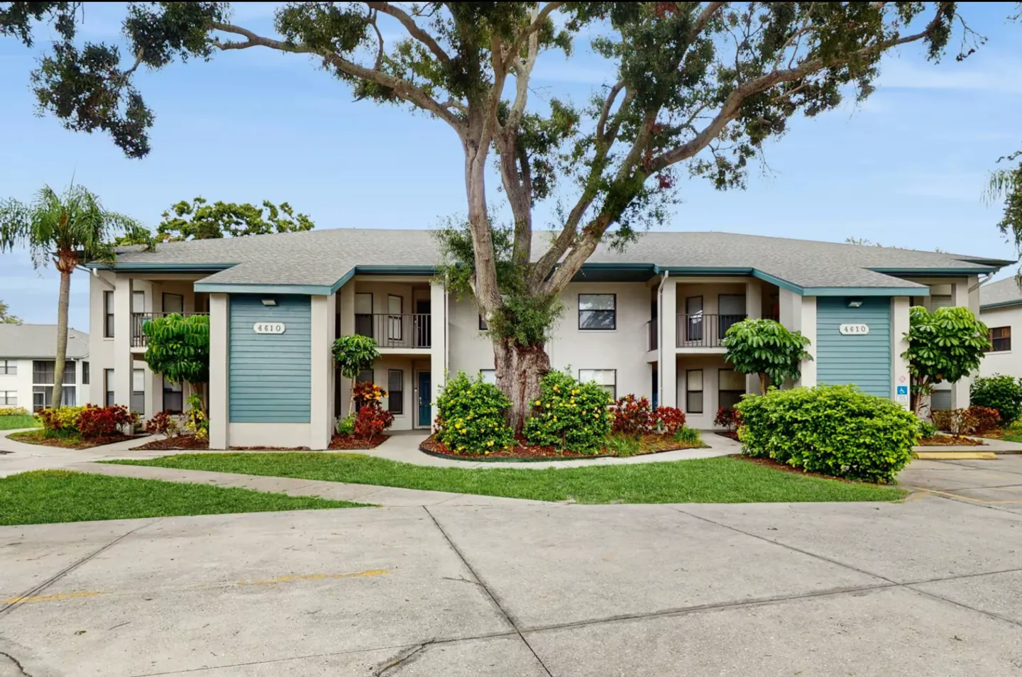 Exterior view of a two-story apartment building with teal siding accents, surrounded by landscaped bushes and a large central tree.