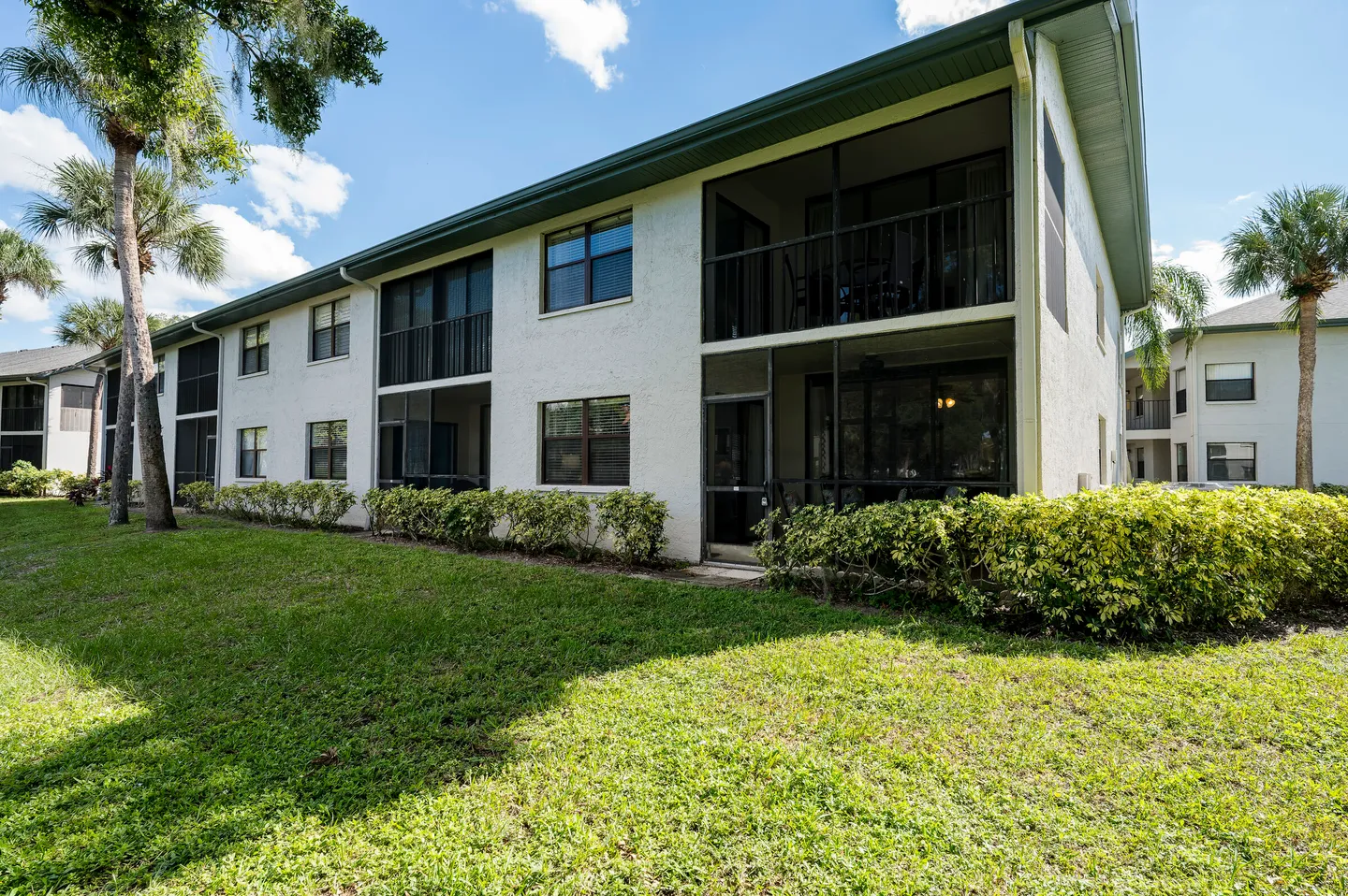 Exterior view of a two-story multi-unit residential building with screened-in porches, manicured bushes, and a green grass lawn.