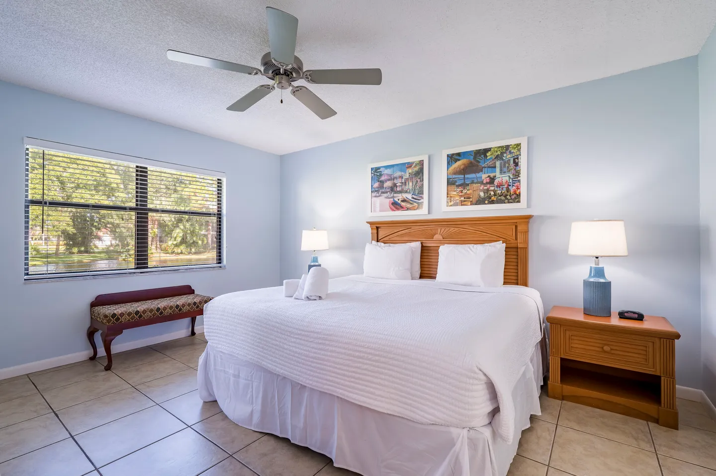 Bedroom with light blue walls, tiled floor, a bed with a wooden headboard, two nightstands with lamps, and a window with blinds overlooking water.
