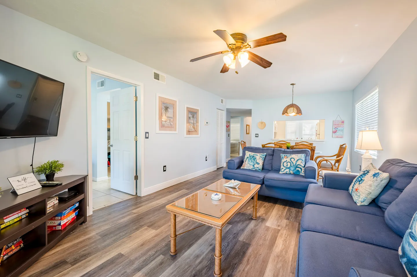 Living room with blue upholstered sofas, wood-look flooring, a wall-mounted TV, and an open dining area in the background.