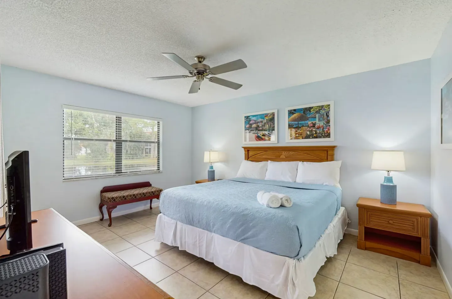 Bedroom with tile flooring, a large bed with blue bedding, matching wooden nightstands with lamps, and a window.