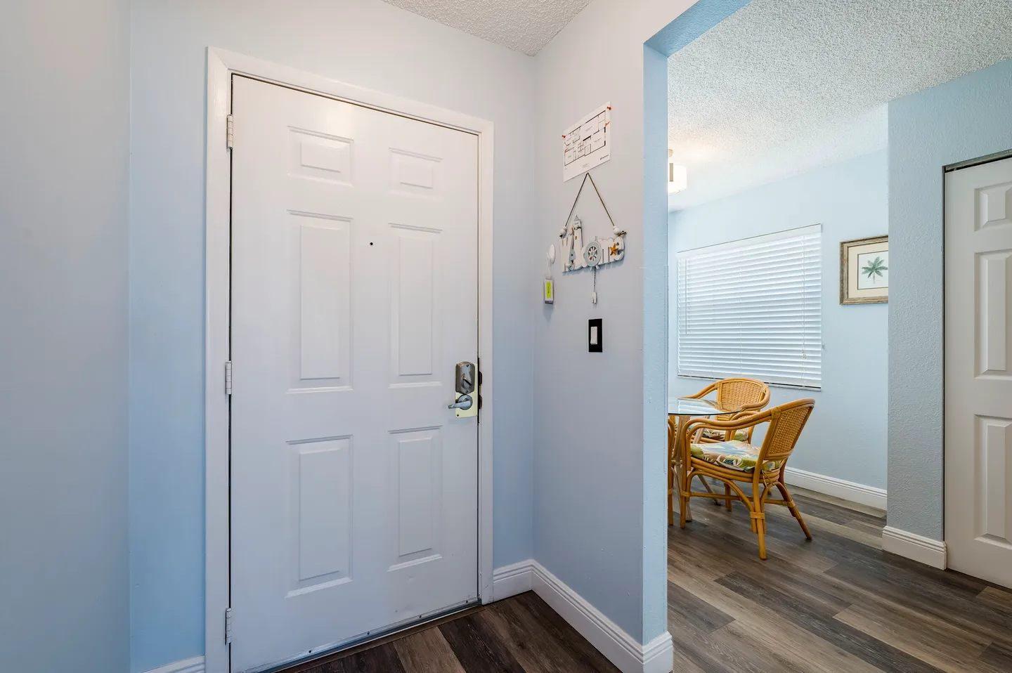 Entryway with a white front door and light blue walls leading into a dining area with wicker chairs and wood-look flooring.