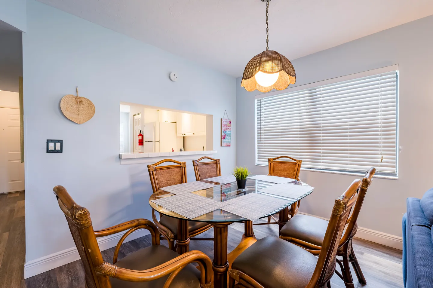 Dining area with a round glass-top table, wicker-style chairs, an overhead pendant light, and a pass-through window to the kitchen.