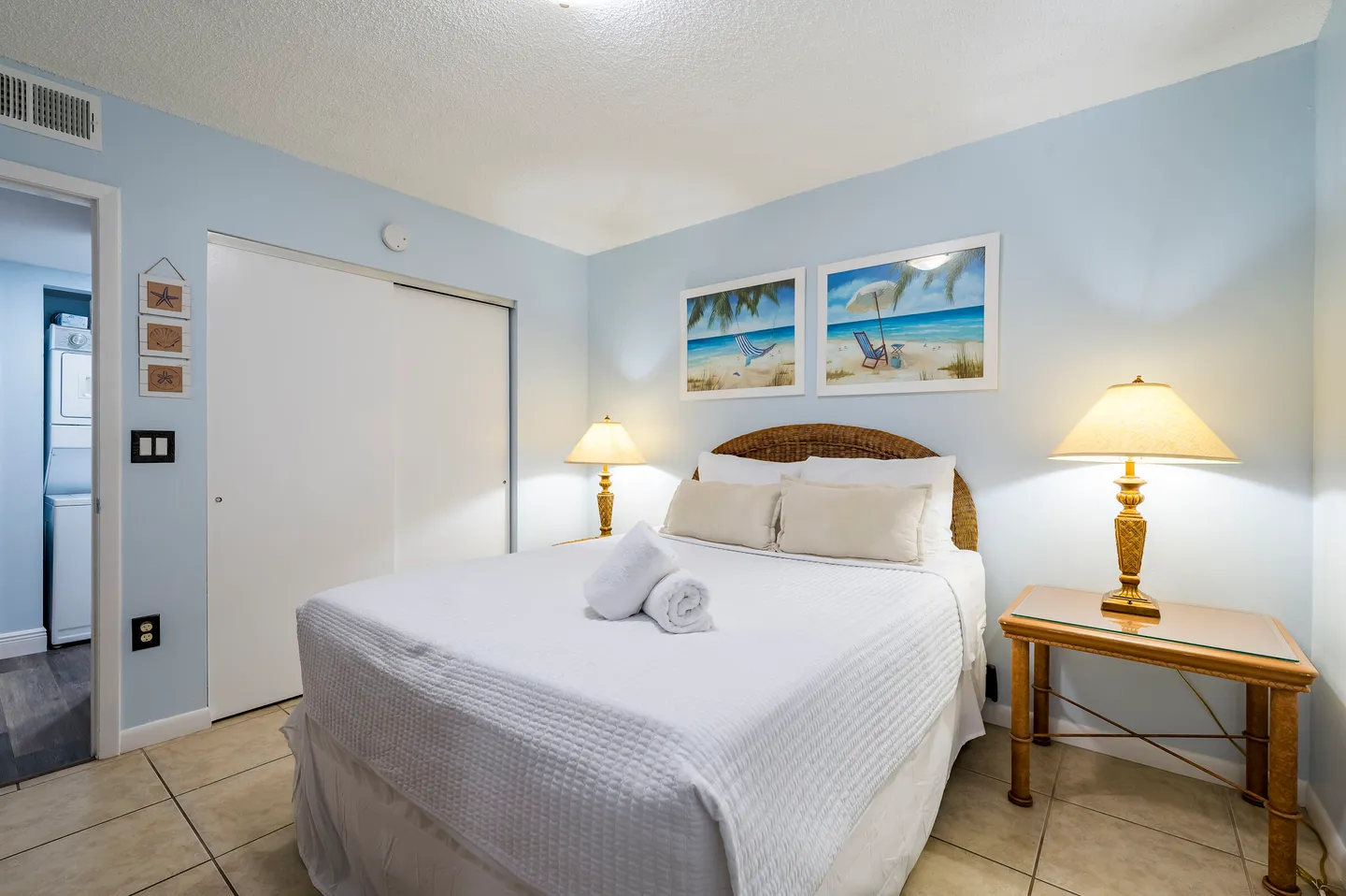 Bedroom with light blue walls, a bed made with white linens, two lamps on nightstands, tiled floors, and framed beach art.