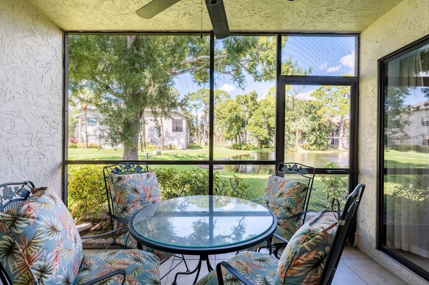 Screened porch with a round glass-topped table and cushioned chairs overlooking trees and a pond.