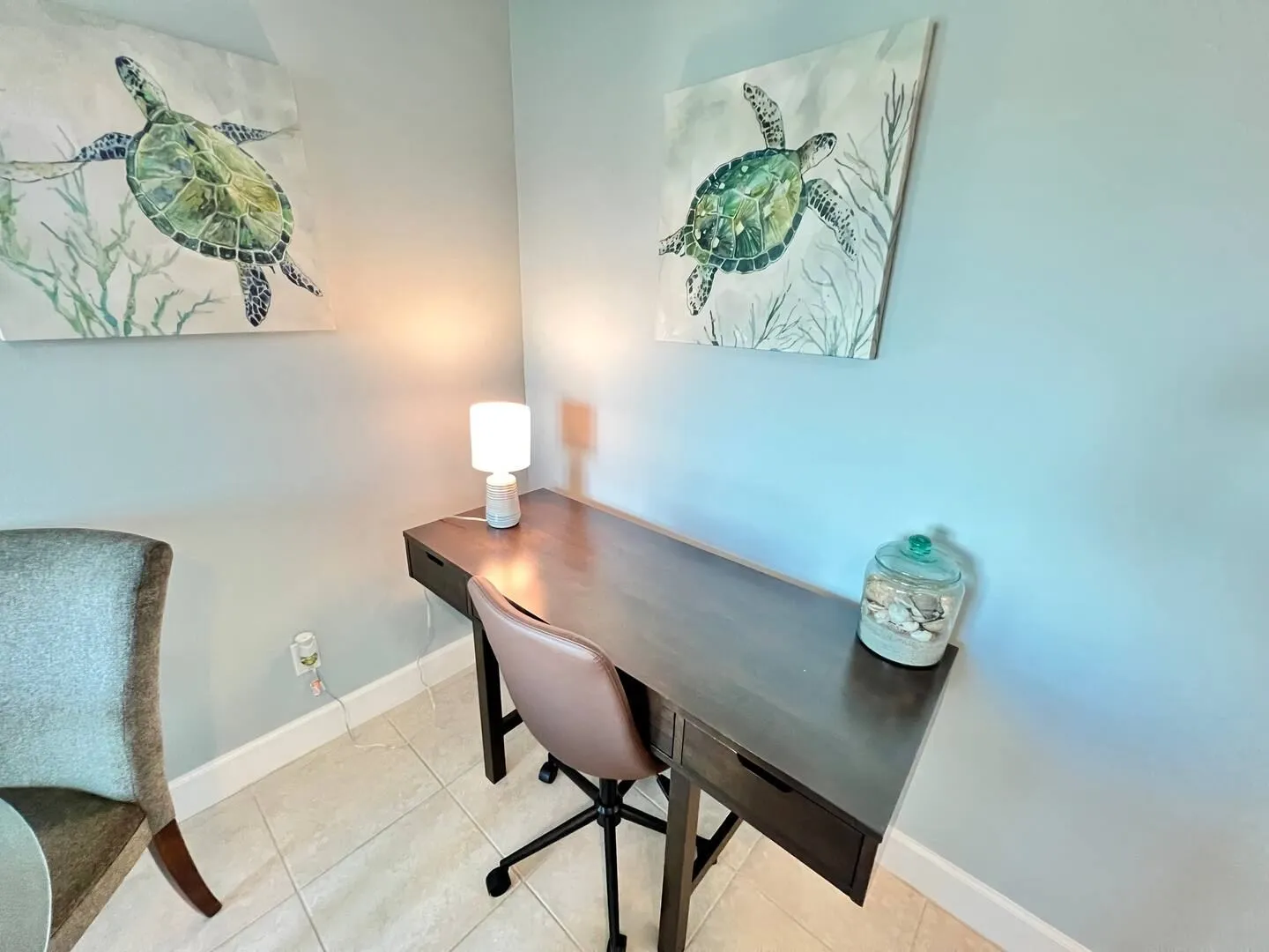 A dedicated workstation tucked into a corner with a dark wood desk, rolling office chair, small lamp, and sea turtle wall art.