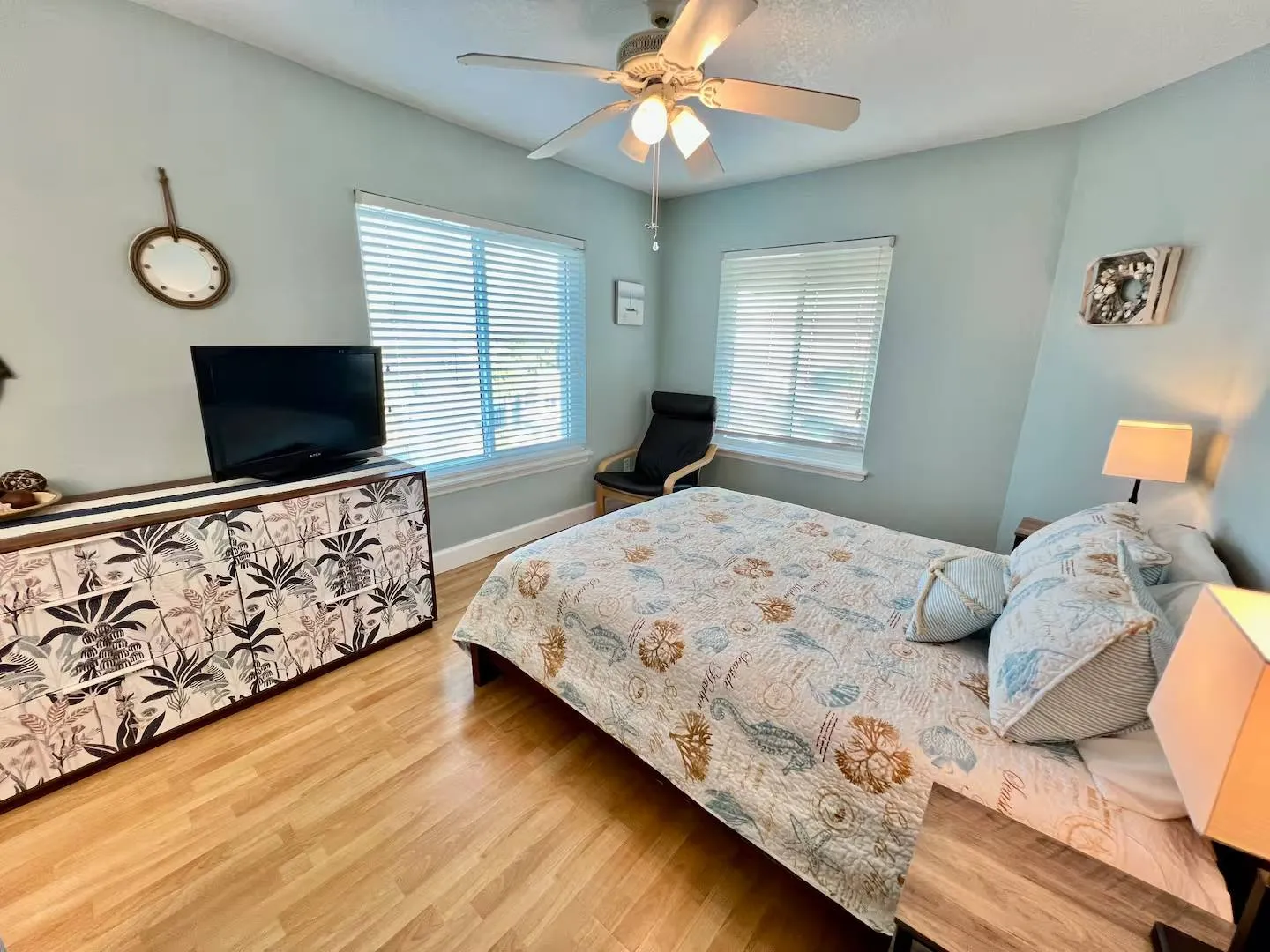 Bedroom with coastal-themed bedding, a flat-screen TV on a tropical-patterned dresser, light wood-look floors, and light blue walls.