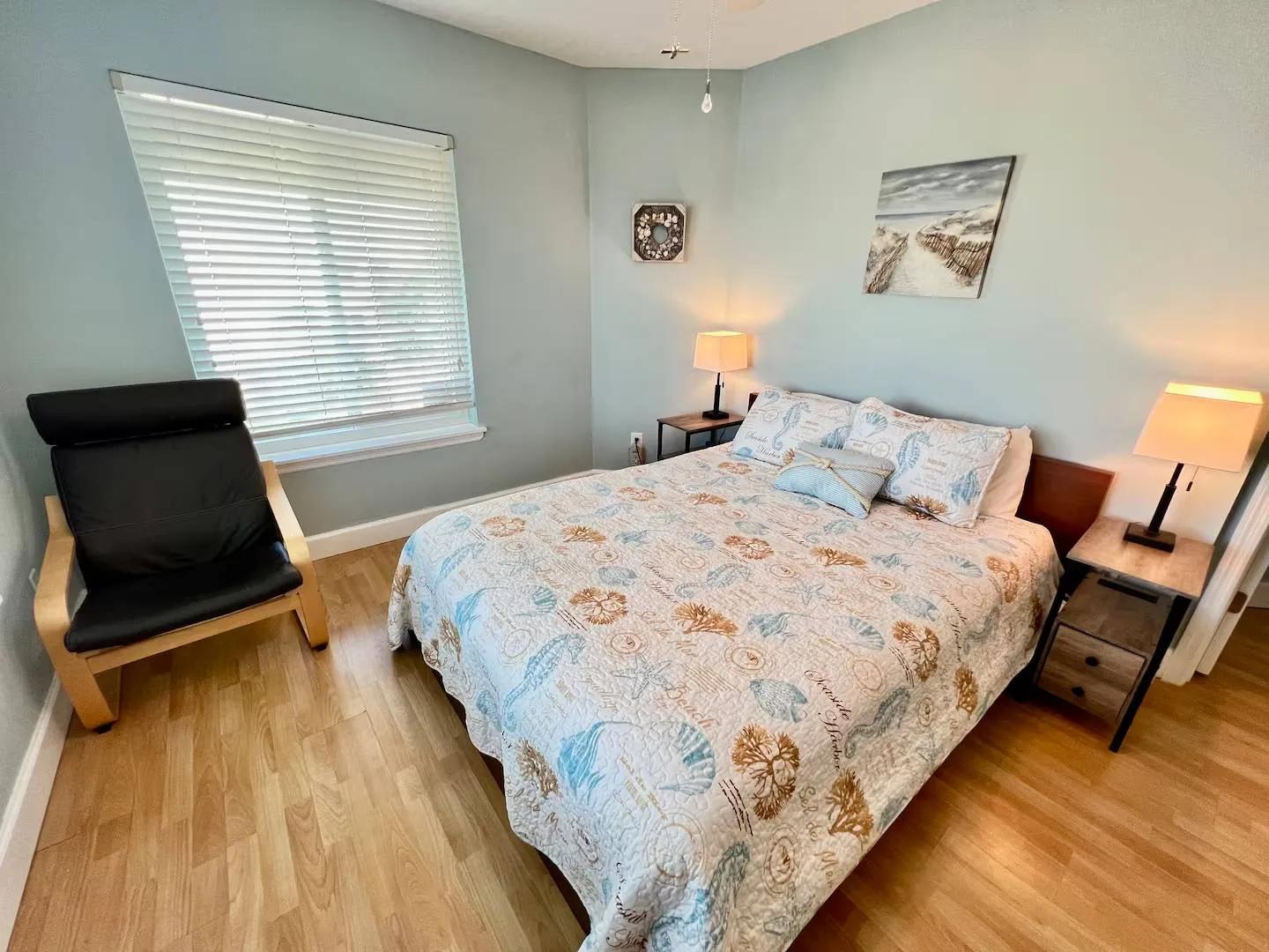 Bedroom with wood-look flooring, light blue walls, a bed made with a coastal-patterned quilt, two nightstands with lamps, a window with horizontal blinds, and an armchair.
