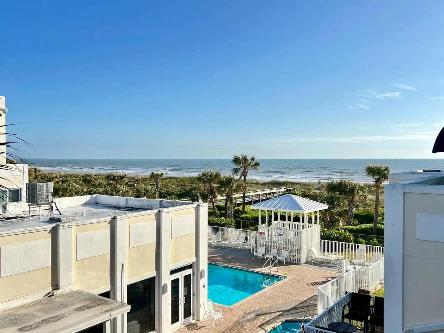 Elevated view overlooking an outdoor swimming pool, gazebo, and dunes leading to the ocean under a clear blue sky.