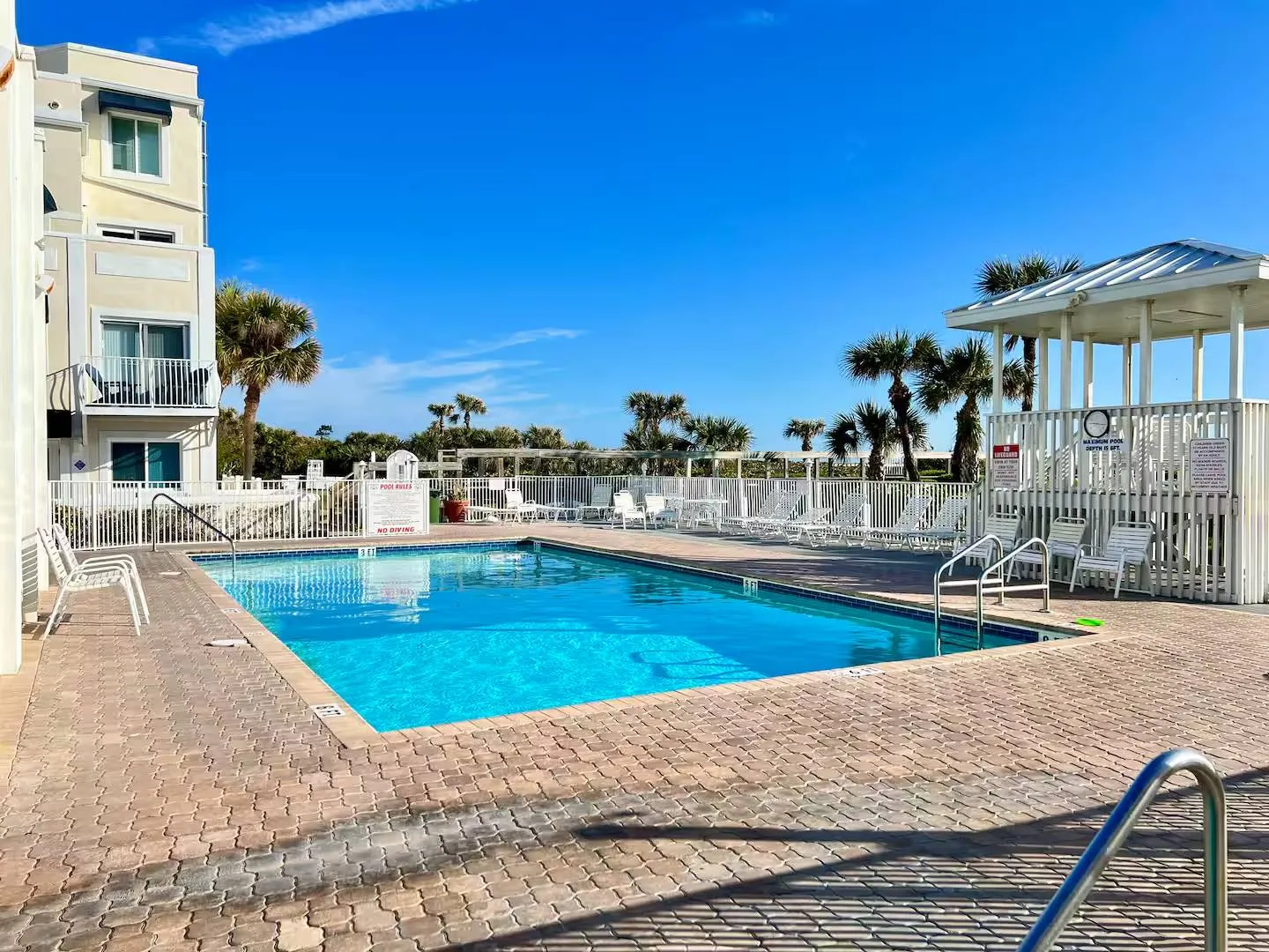 An outdoor rectangular swimming pool surrounded by a brick paver deck, white lounge chairs, a covered gazebo, and a multistory residential building under a bright blue sky.