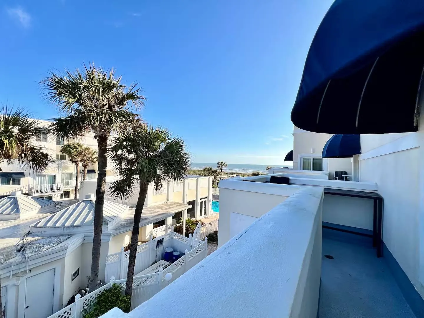 Balcony view overlooking nearby coastal buildings, palm trees, a swimming pool, and the ocean under a clear blue sky.