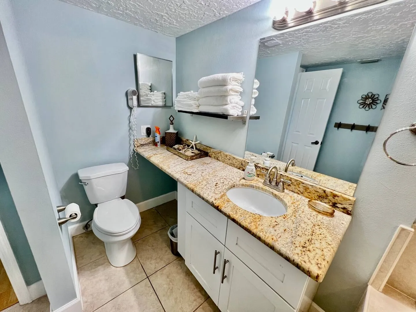 Bathroom featuring a white vanity with a speckled countertop, an undermount sink, a large mirror, a toilet, and light blue walls.