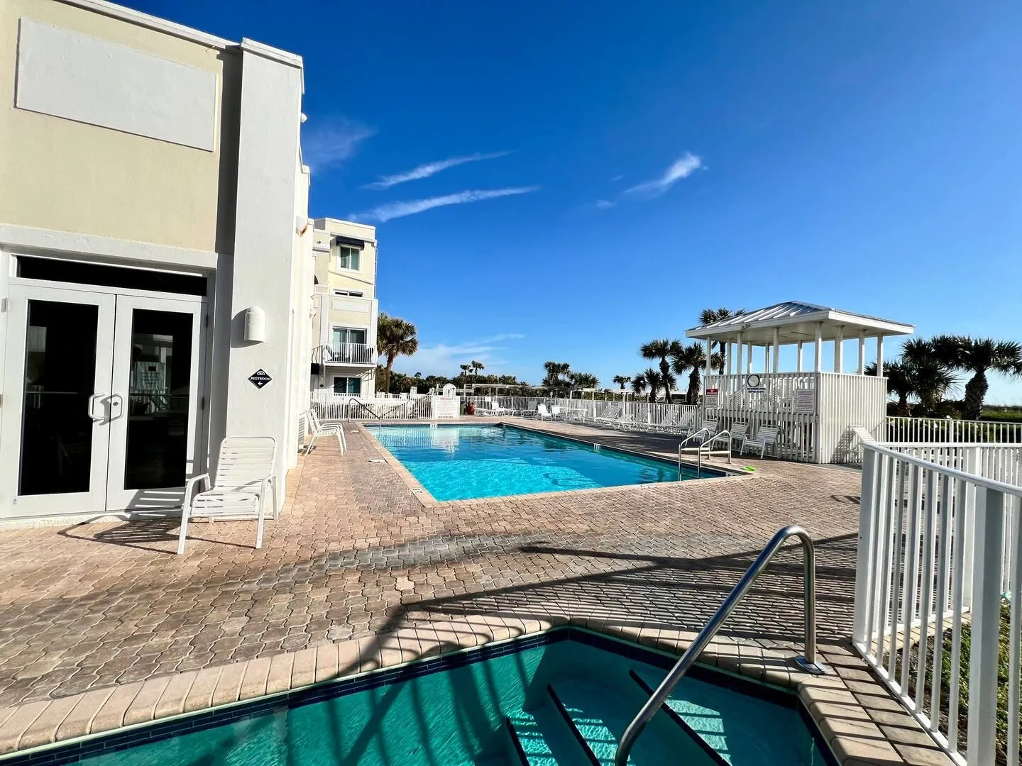 Outdoor swimming pool area and hot tub surrounded by a brick paver patio next to a multi-story building and white gazebo.