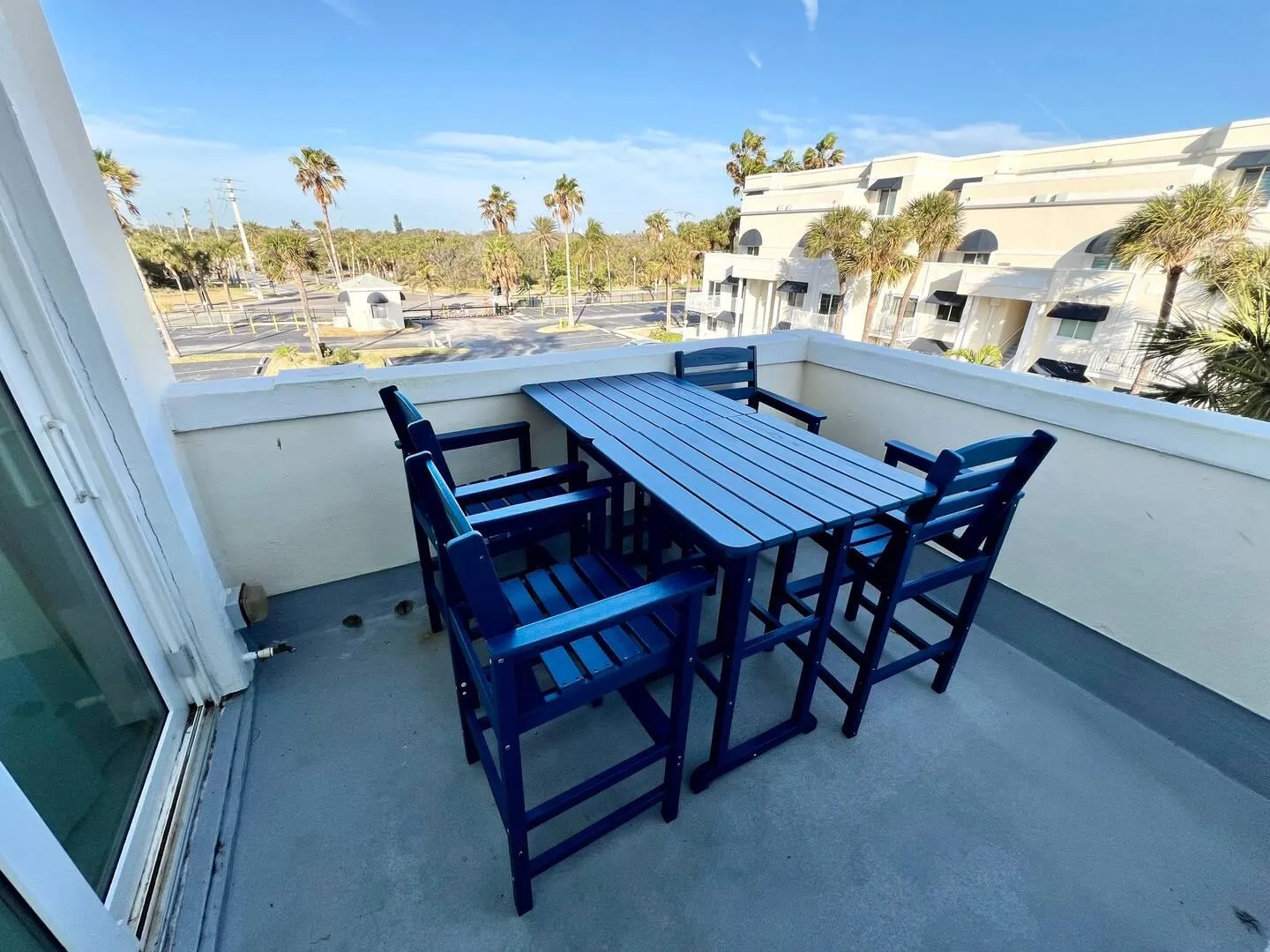Balcony with a blue slatted dining table and high chairs overlooking palm trees, surrounding buildings, and a clear blue sky.