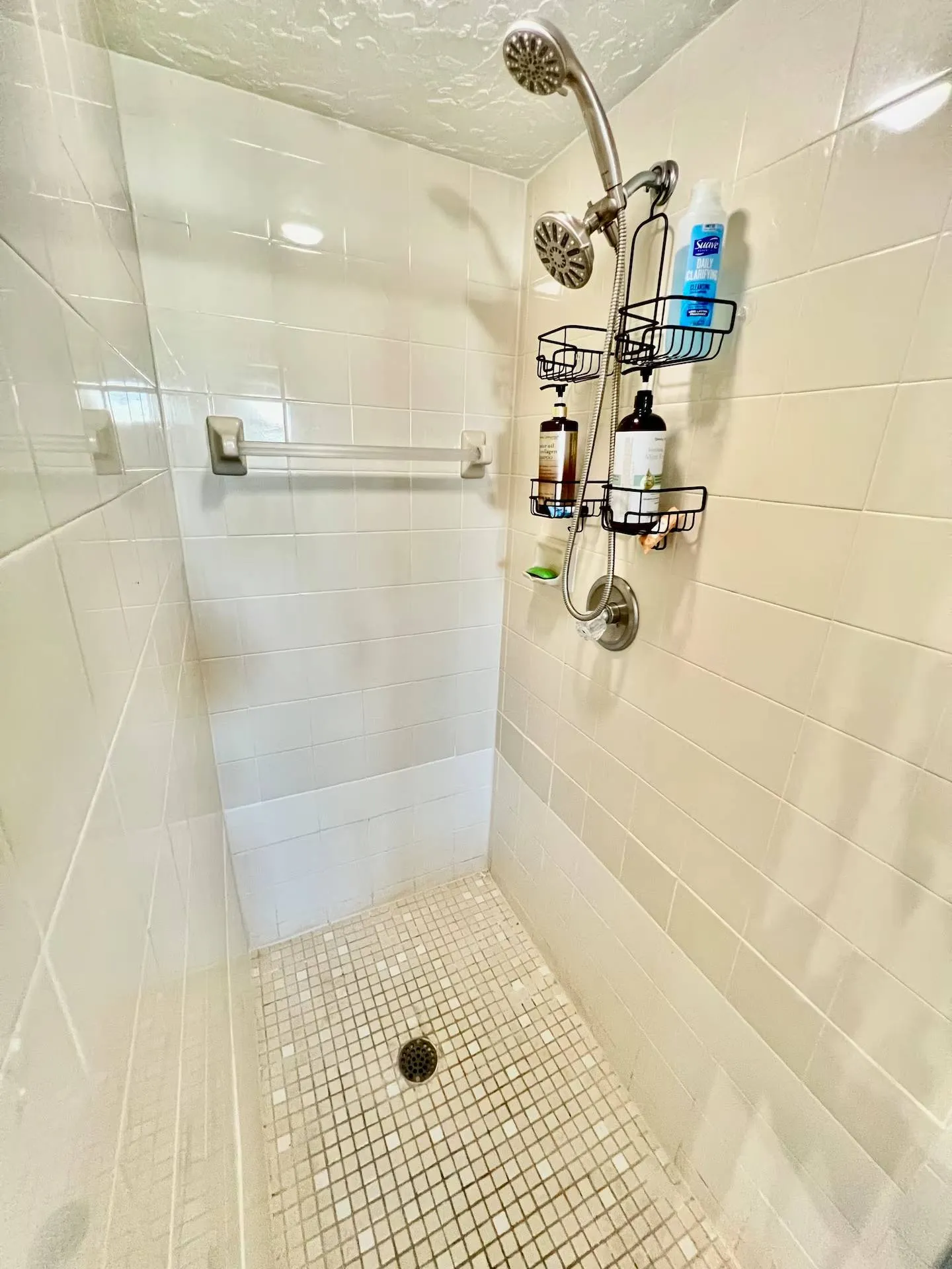Walk-in shower with square tiled walls, a mosaic tile floor, glass door, and a metal shower caddy holding toiletries.