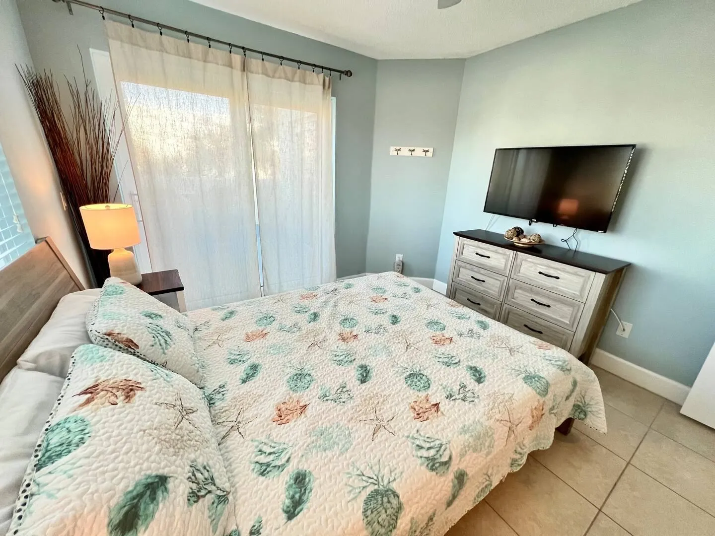 Bedroom with coastal seashell bedding, tile floors, a dresser with a wall-mounted TV, and sliding glass doors covered by sheer curtains.