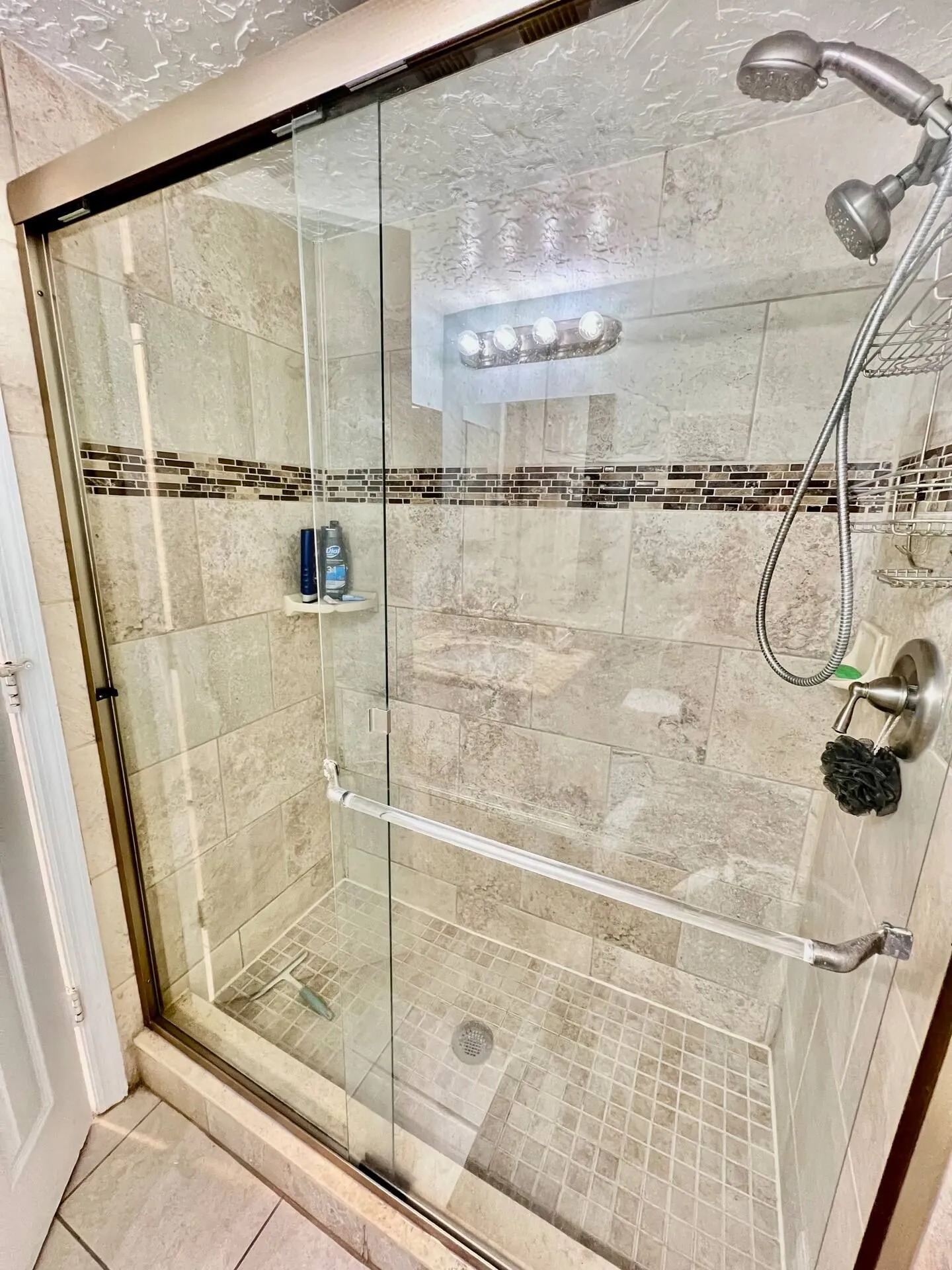 Walk-in shower with sliding glass doors, neutral stone-look wall tiles with a mosaic accent border, and a handheld showerhead.