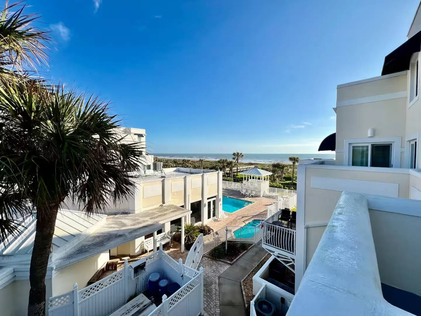 Elevated view from a balcony showing surrounding resort buildings, swimming pools, palm trees, and the ocean under a clear blue sky.