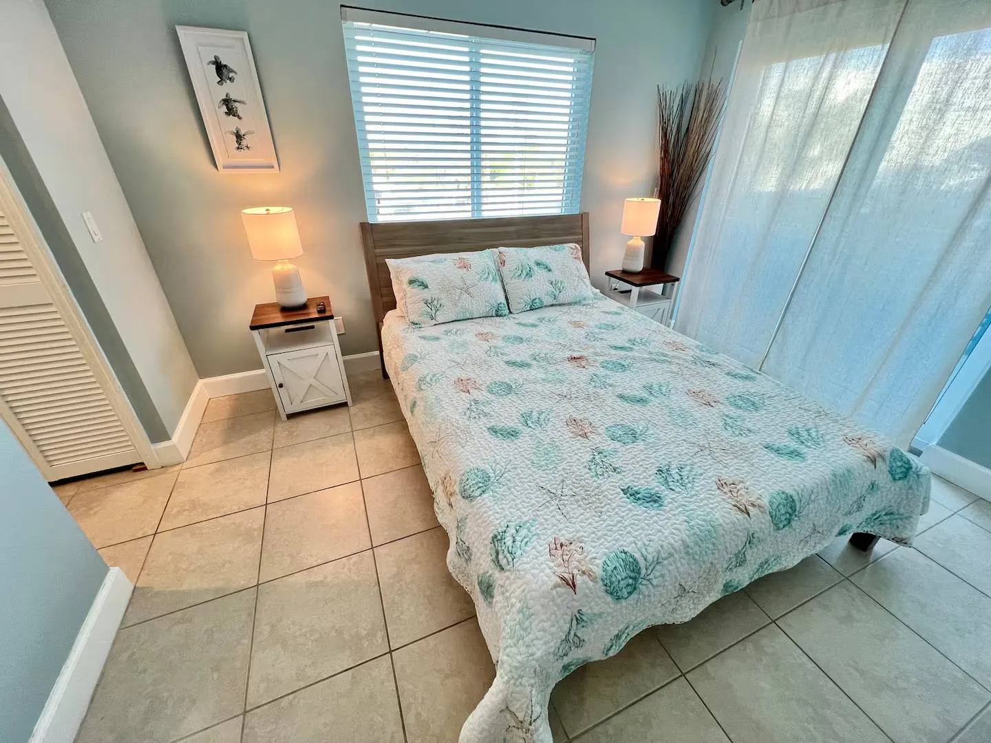 Bedroom with a bed dressed in coastal-themed bedding, tiled floors, bedside tables with lamps, and a window with blinds.