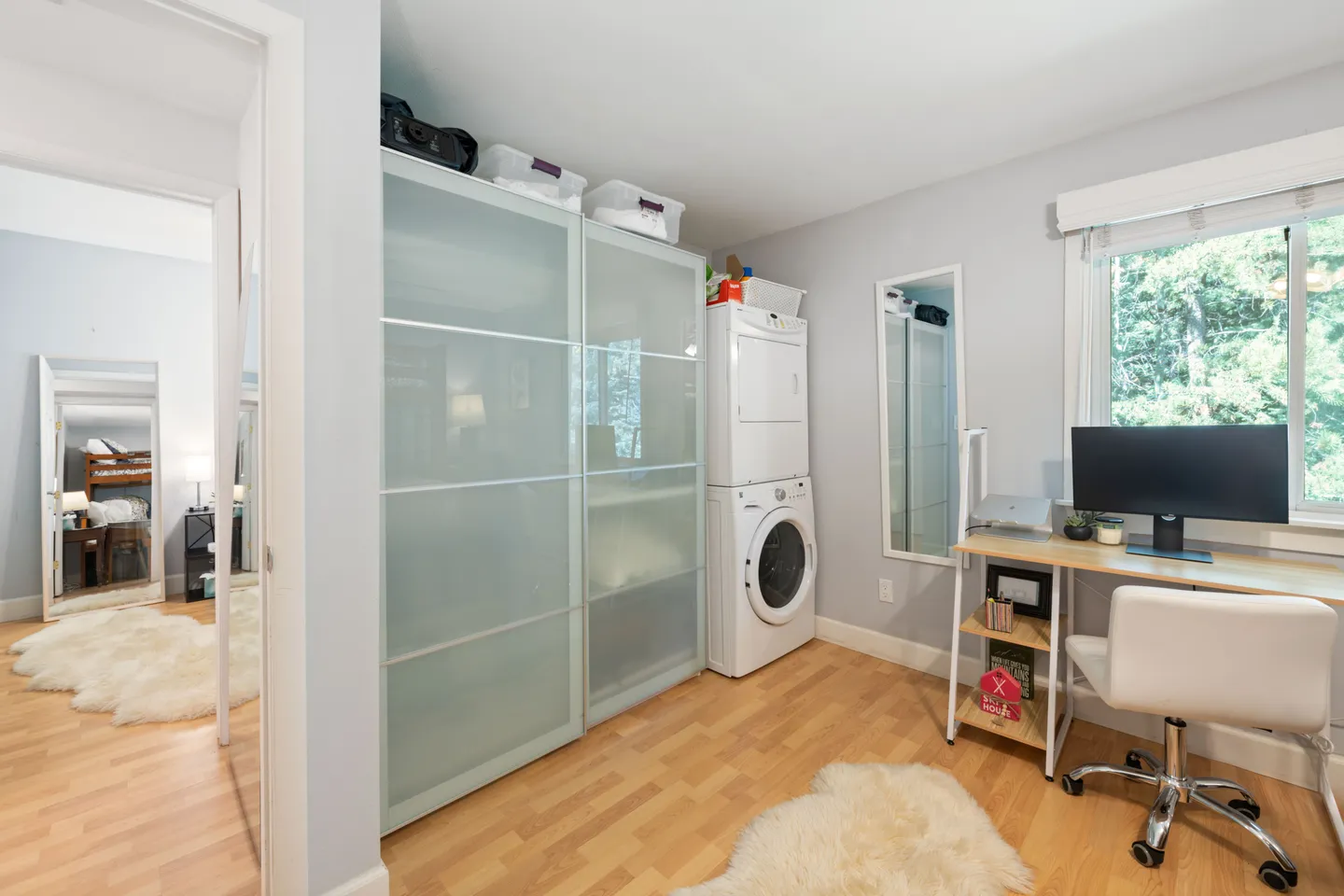 A multipurpose room with a desk and computer, stacked washer and dryer, large frosted glass sliding wardrobe, and light wood-look floors.