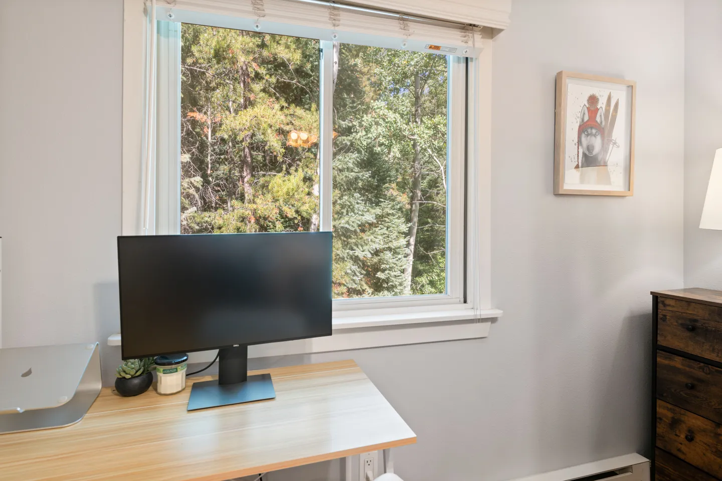 A light wood desk with a computer monitor set below a window overlooking evergreen trees.