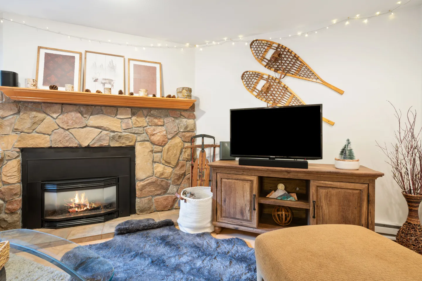 Living room corner featuring a stone fireplace with lit fire, wood mantel, television on a wooden media console, and decorative vintage snowshoes on the wall.