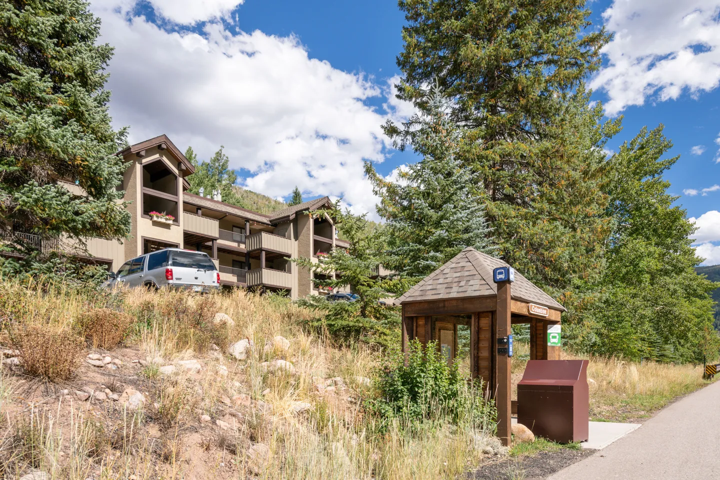 Exterior view of a multi-story condo complex on a grassy hill, with a wooden bus stop shelter in the foreground.