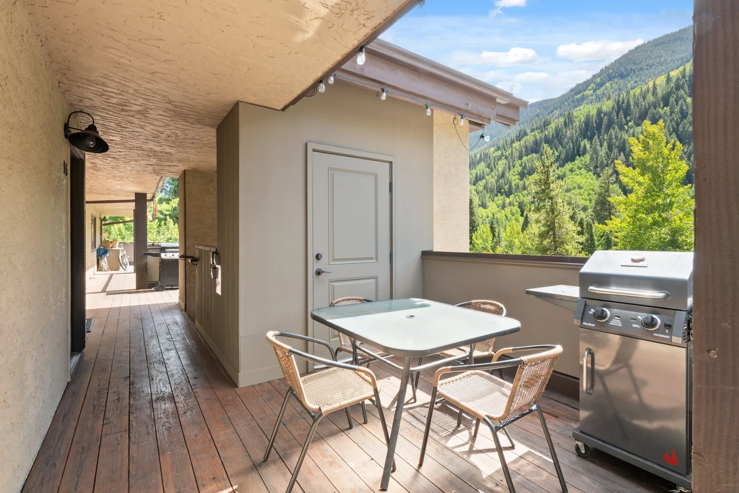 Outdoor covered balcony with a dining table, chairs, barbecue grill, and view of forested mountains.