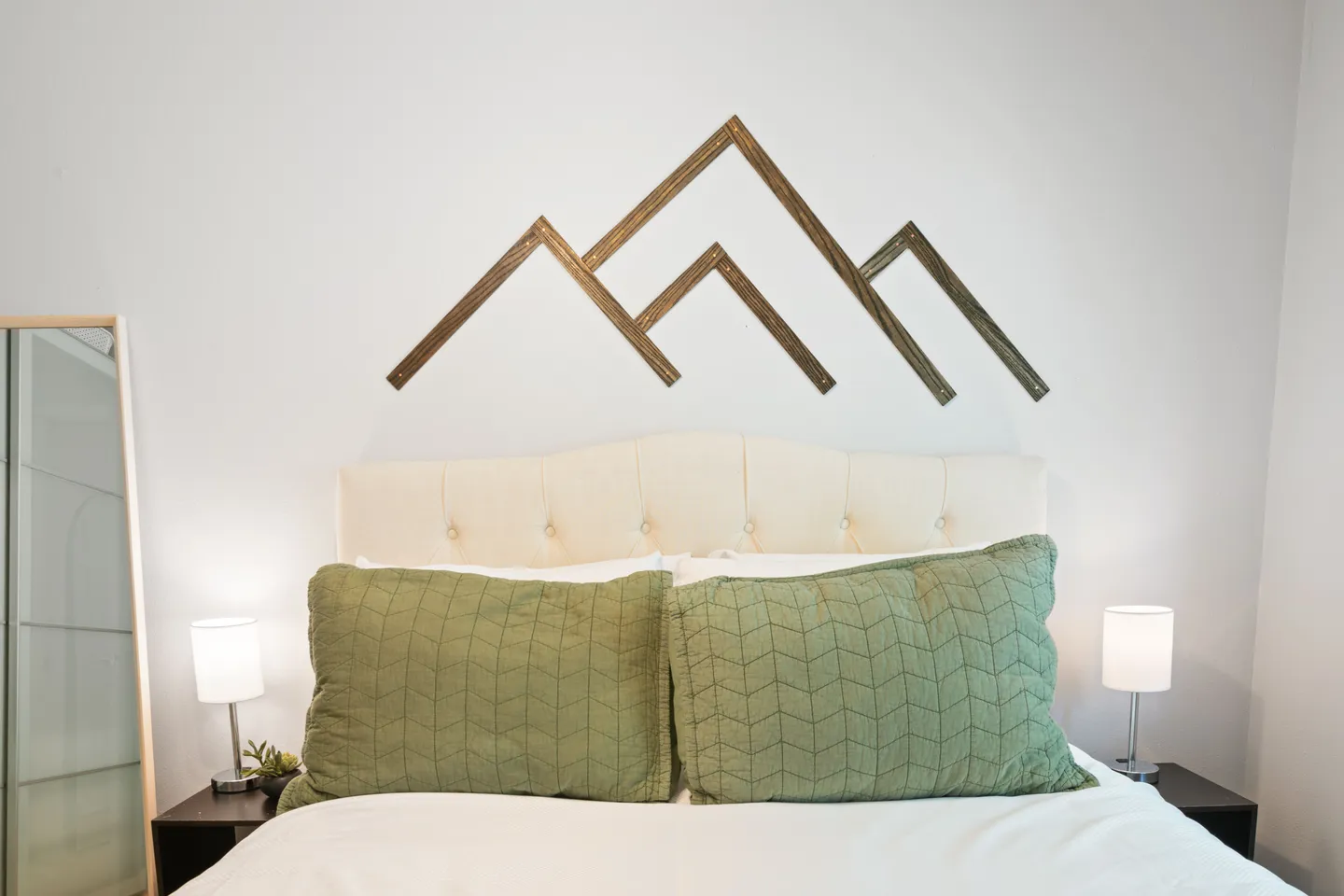 Bedroom headboard with tufted cream upholstery, two green pillows, bedside lamps, and wooden geometric mountain wall art.