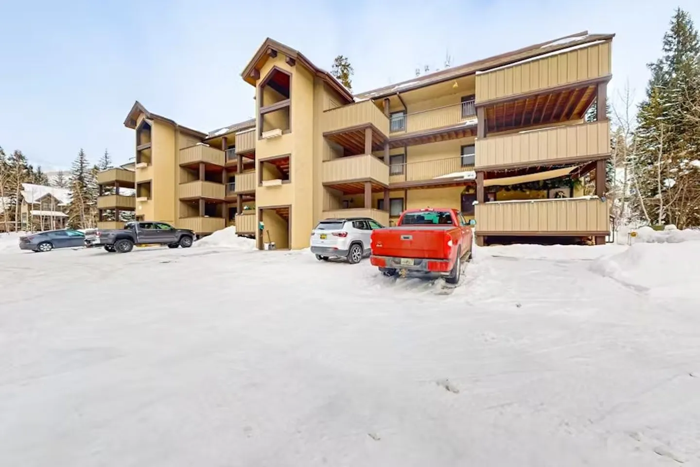 Exterior view of a multi-story tan condominium building with balconies and a snow-covered parking area in front.