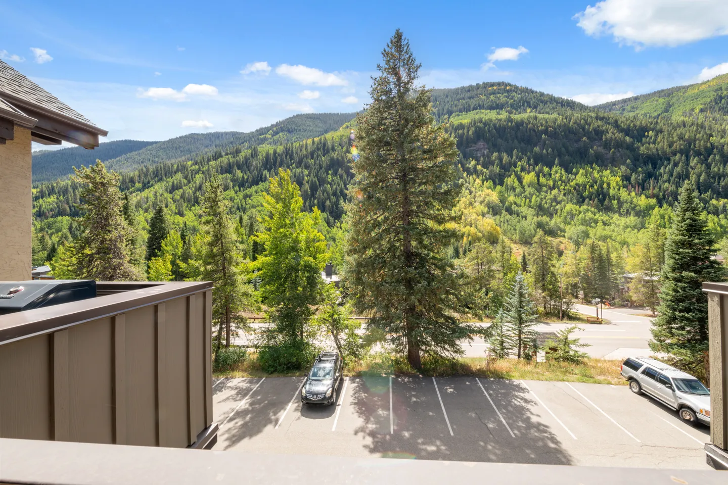 Balcony view overlooking a parking lot with parked cars, dense evergreen trees, and forested mountains under a blue sky.