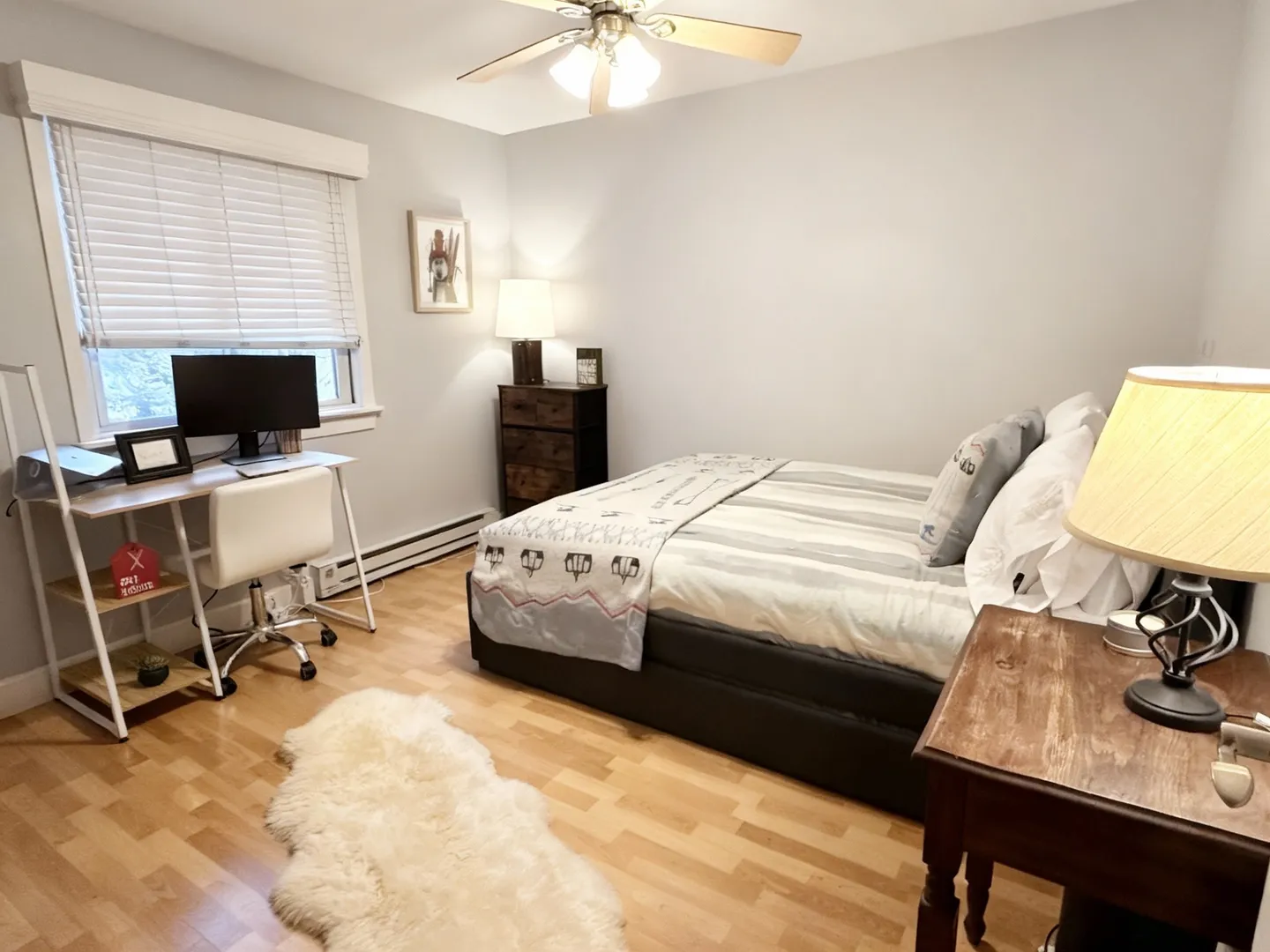 Bedroom featuring a made bed, wood-finish flooring, a desk with a monitor and chair, and a ceiling fan.