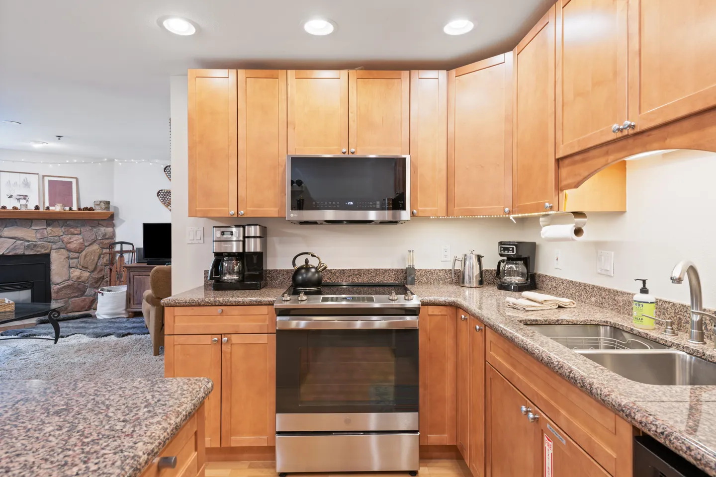 Kitchen with light wood shaker cabinets, granite countertops, stainless steel appliances, and a view into the living room with a stone fireplace.