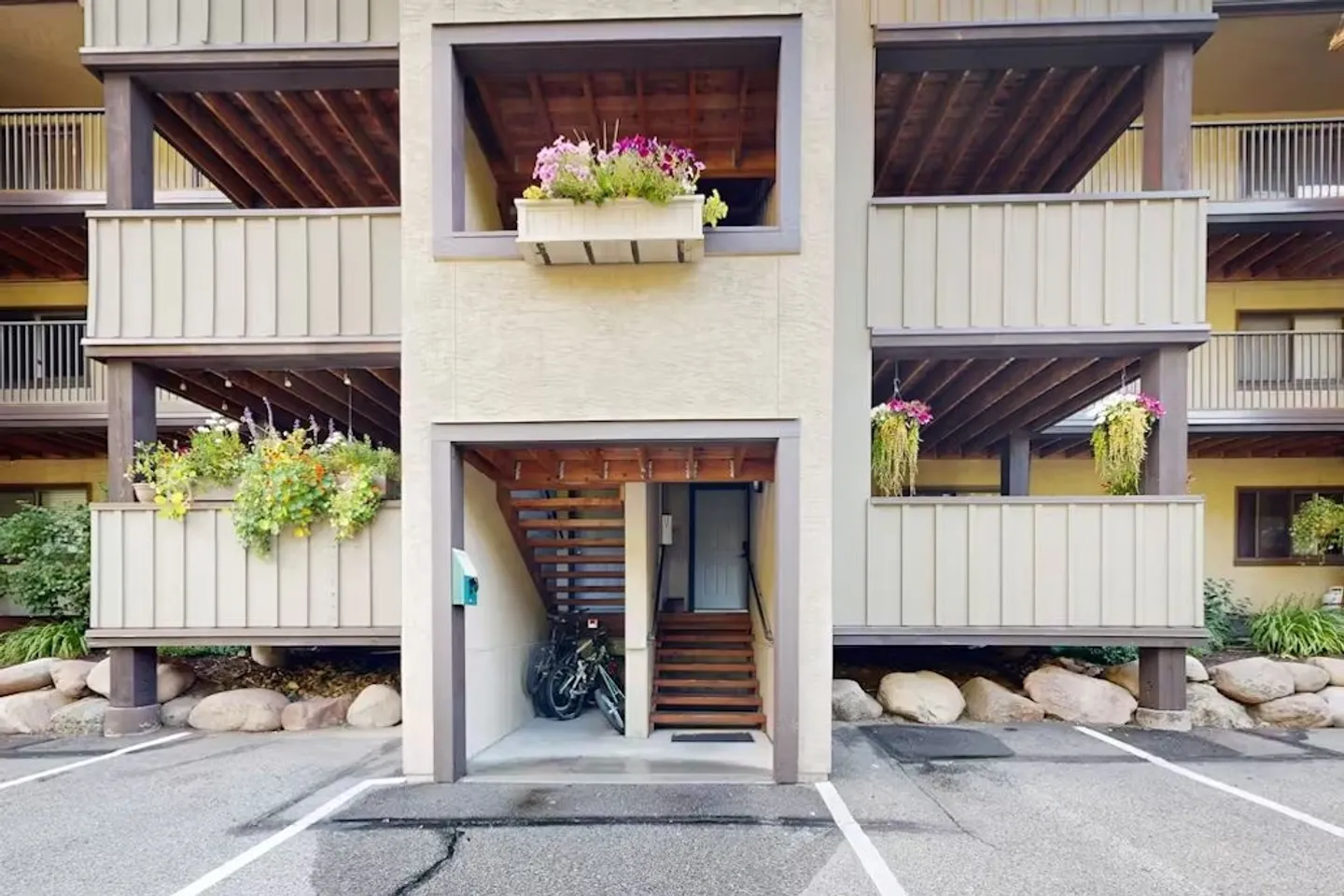 Exterior of a multi-story building featuring wooden balconies, outdoor stairs leading to an entrance, and parking spaces in front.