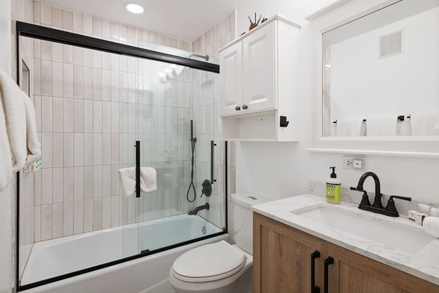 Bathroom with a shower-tub combination featuring sliding glass doors with black trim, a toilet, a light wood vanity, and a white upper cabinet.