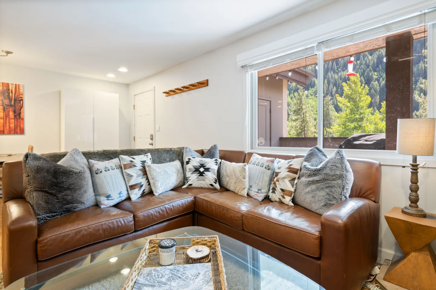 Living room with a brown leather sectional sofa adorned with patterned pillows, situated next to a large window overlooking trees.
