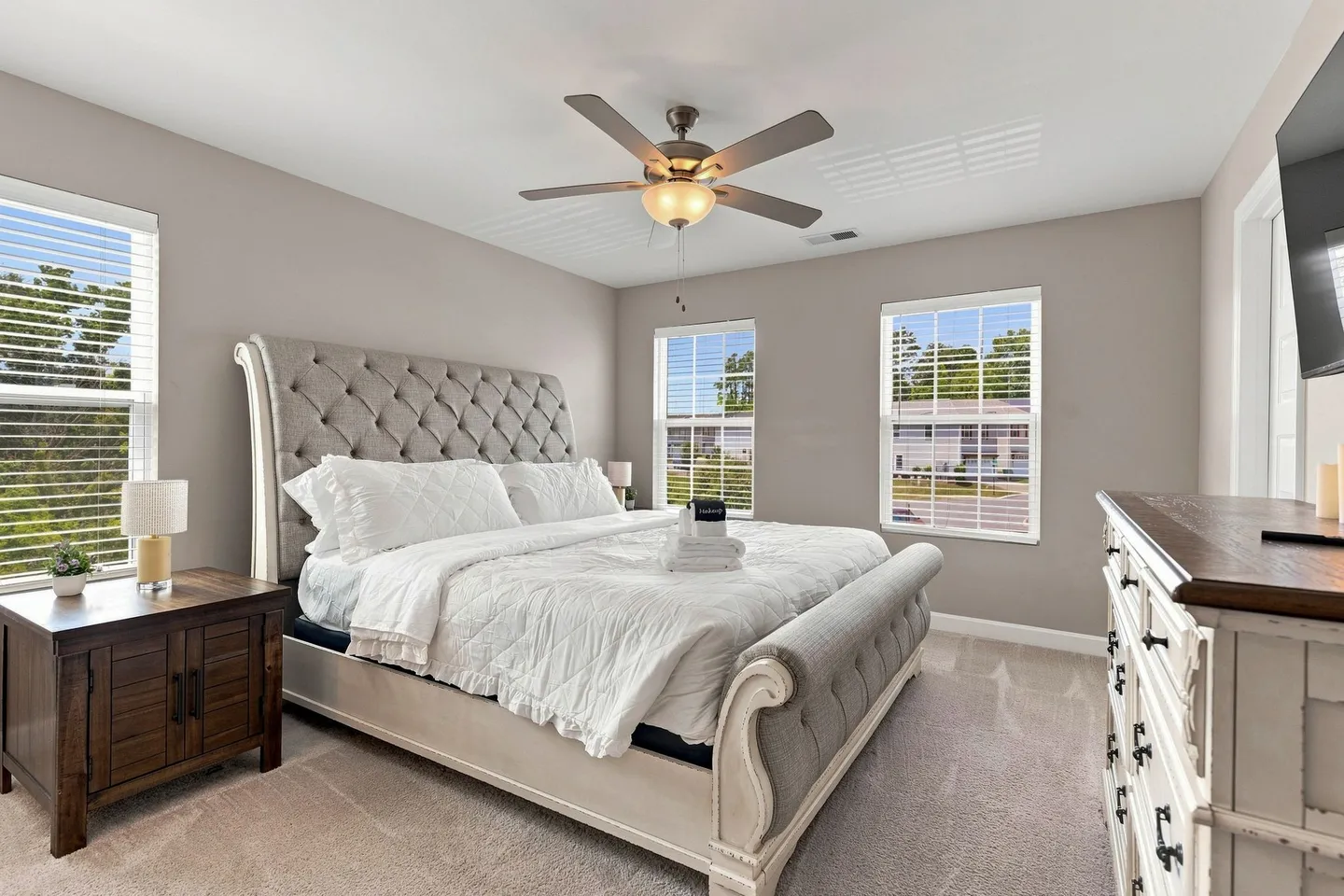 Bedroom with a tufted sleigh bed, multiple windows, carpeted floor, ceiling fan, and wooden dressers.