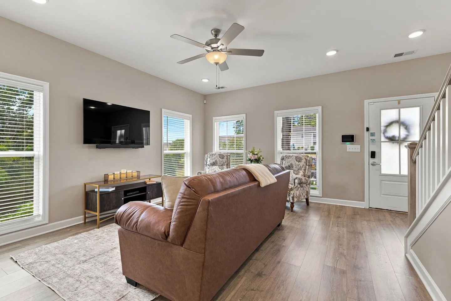 Living room featuring a brown sofa, a wall-mounted TV over a media console, armchairs, and an entry door next to stairs.