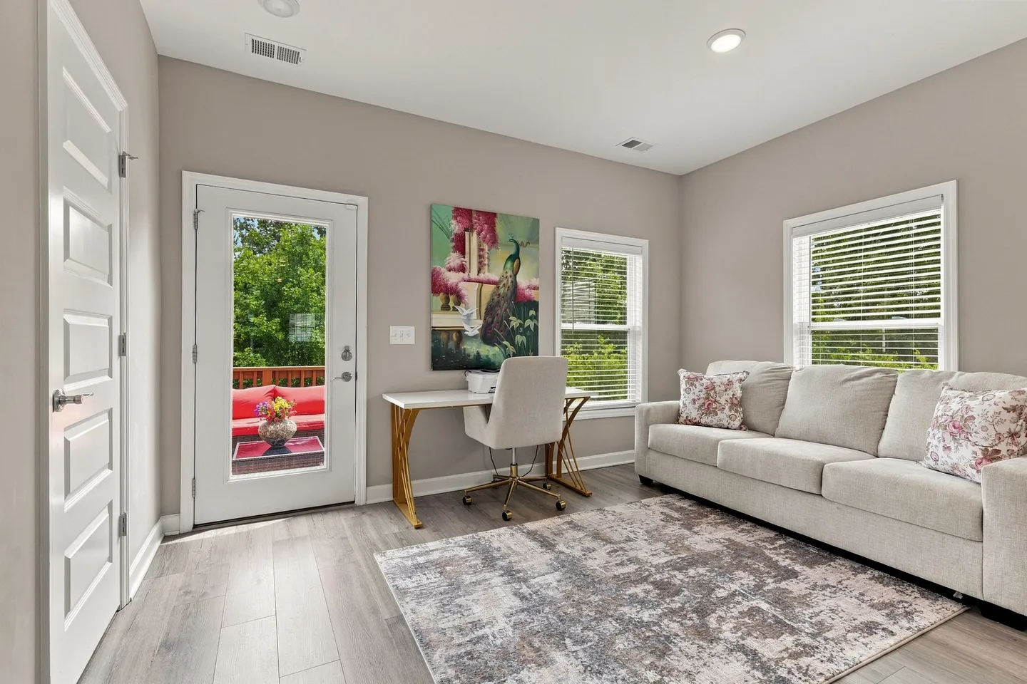 Bright room with wood-style flooring, a light-colored sofa, an office desk with chair, and a glass-paneled exterior door leading to a deck.